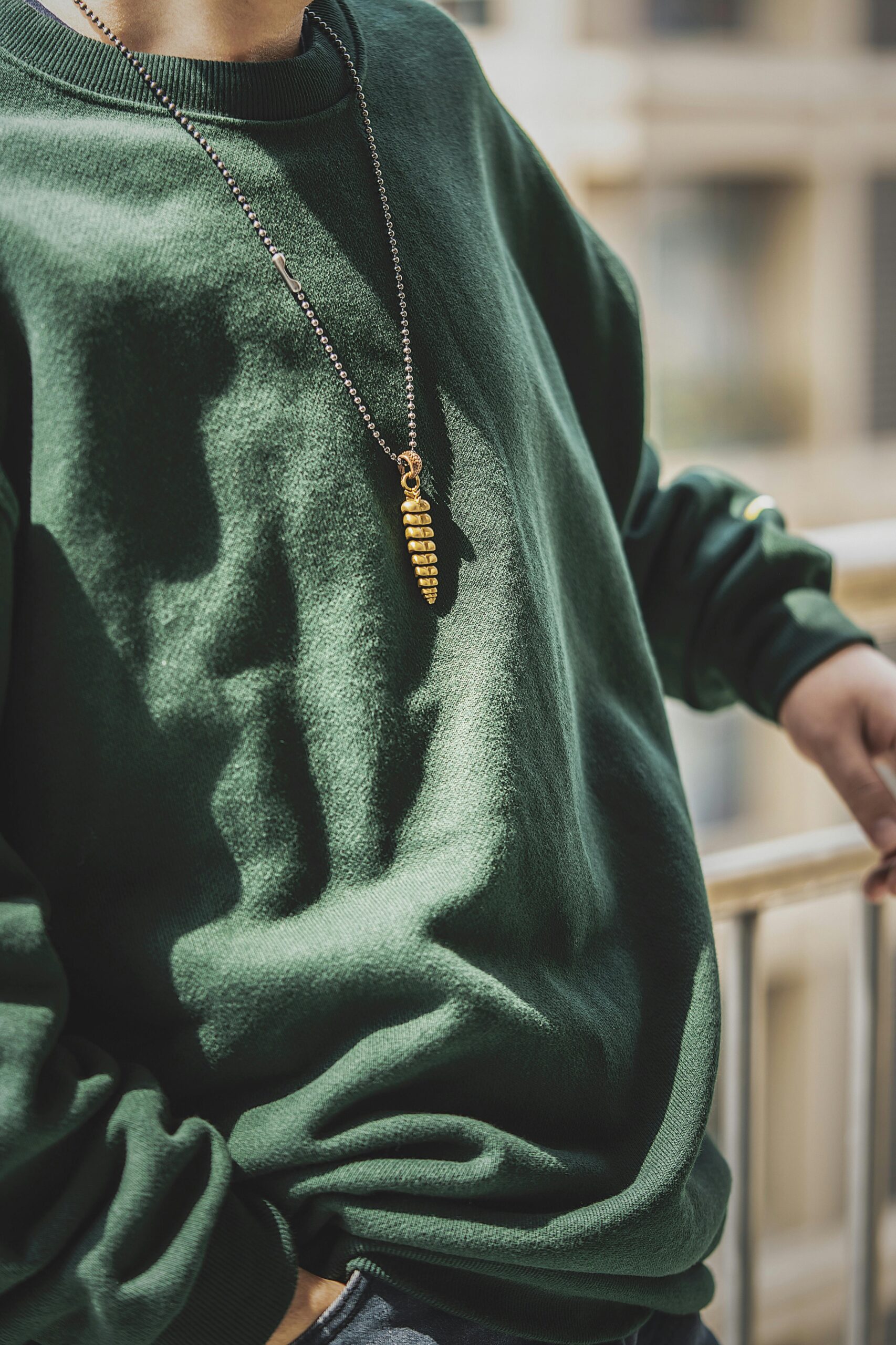 A stylish close-up of a person wearing a green sweatshirt with a unique gold pendant necklace, captured outdoors.