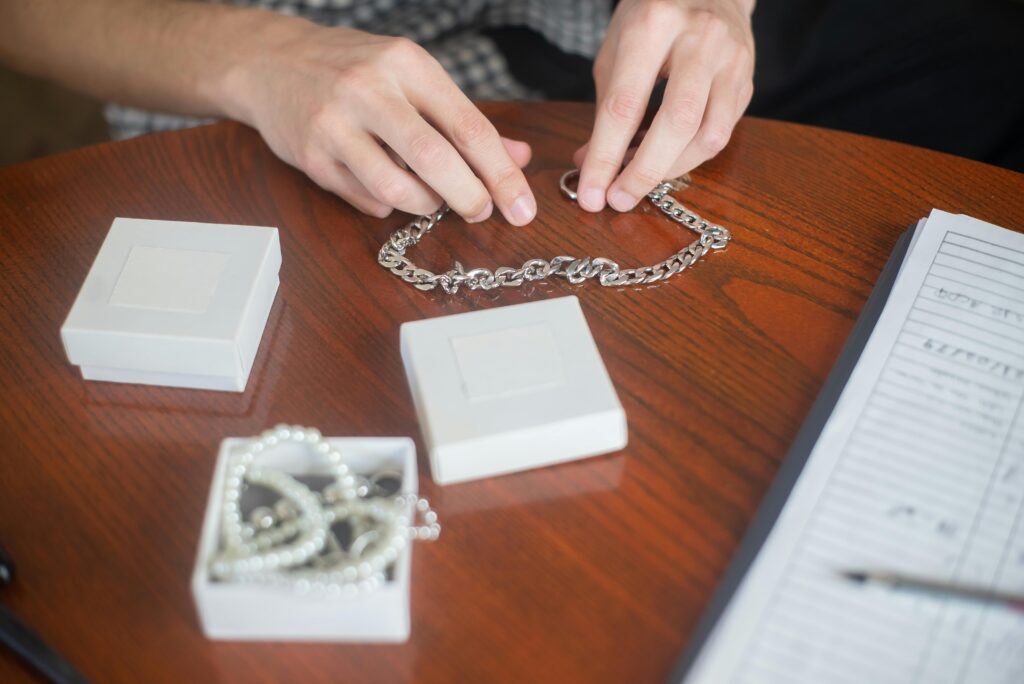 Hands arranging a silver chain necklace on a wooden table with jewelry boxes, indoors.